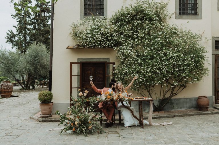 Bride and groom sharing laughs and holding up their glasses as they sit on a wooden table in the middle of the road during their elopement shoot with Alexa Ann Photography