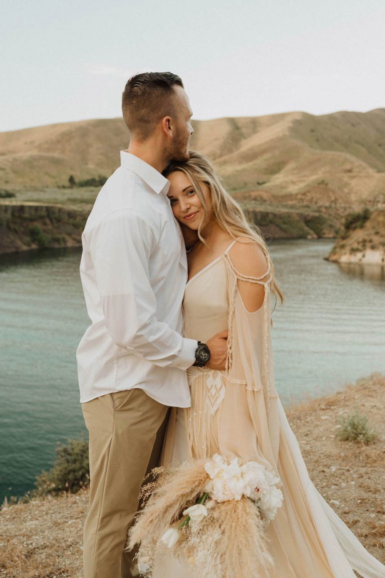 Bride smiles at the camera as she and the groom share an embrace in front of the lake during their elopement shoot with Alexa Ann Photography